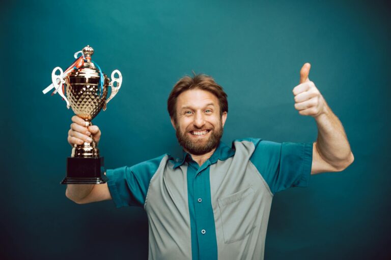 Smiling man holding a trophy and giving a thumbs up gesture against a blue backdrop.