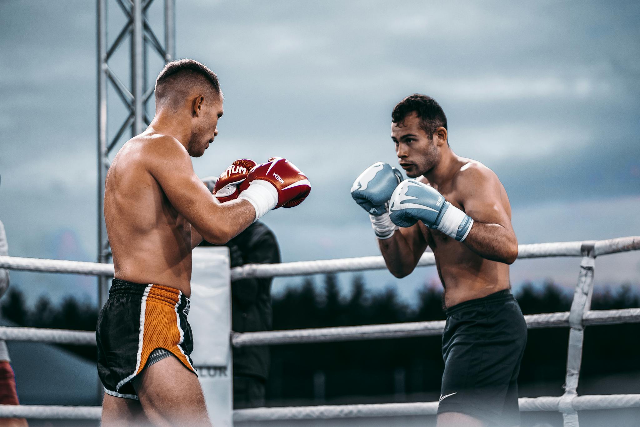 Two male boxers facing off in an outdoor boxing ring under a cloudy sky.